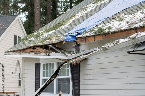 Residential house crushed by fallen trees and tree limbs during severe winter storm with strong winds. Tarp is placed on the damaged rooftop area as a temporary measure before proper roof repairs.