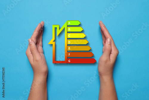 Energy efficiency rating and woman holding hands near it on light blue background, top view photo