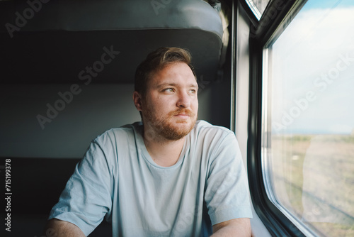 A man with a beard and mustache in a blue t-shirt while traveling by Railway train, sitting in the train and looking out the window.
