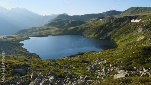 A beautiful blue lake in the mountains. The shore is covered with green grass. Morning, silhouettes of mountain peaks in the background. Cinematic blue mountain lake in a picturesque area.