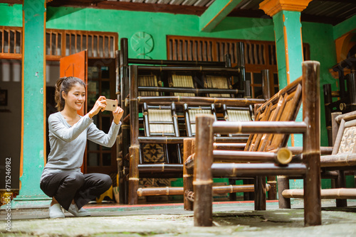 Asian girl squats using her phone camera to take a picture of a bamboo chair at a craft workshop photo