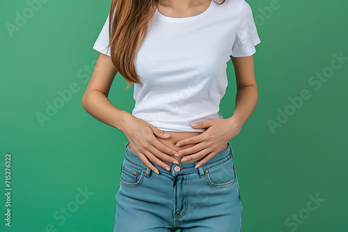 Slender Woman in White T-shirt and Jeans Gently Rest Her Hands on Her Stomach, Embodying a Calm and Centered Presence, Against a Clean and Tranquil Green Background