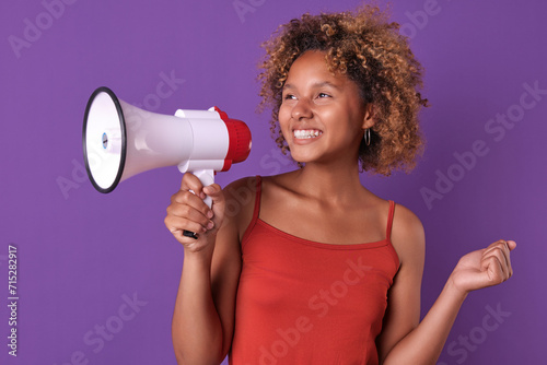 Young happy pretty African American woman teenager smiles widely and holds megaphone in hands rushing to report good news or own achievements in education stands on plain purple background.