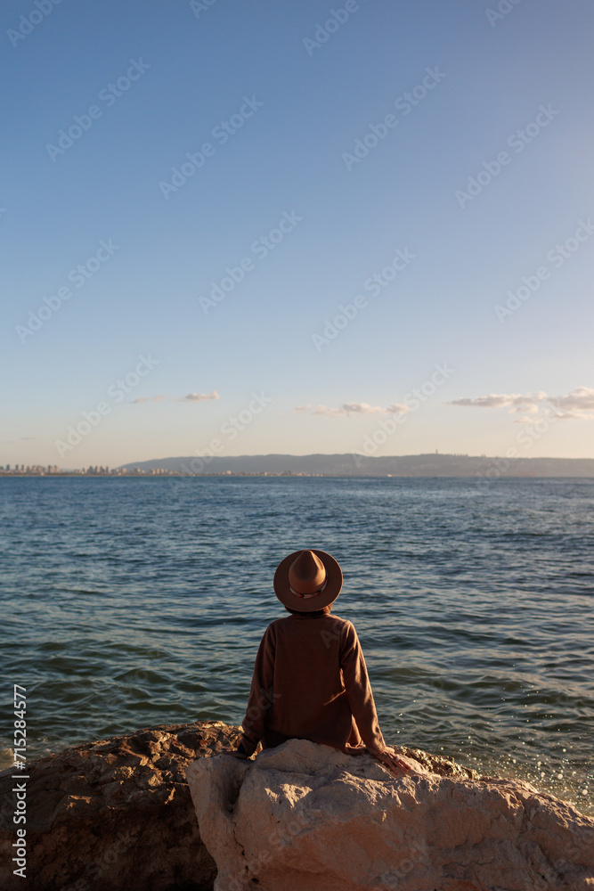 Fototapeta premium back view. a young girl wearing a coat and hat sits on a high stone and looks at the beautiful view of the blue sea and sky.