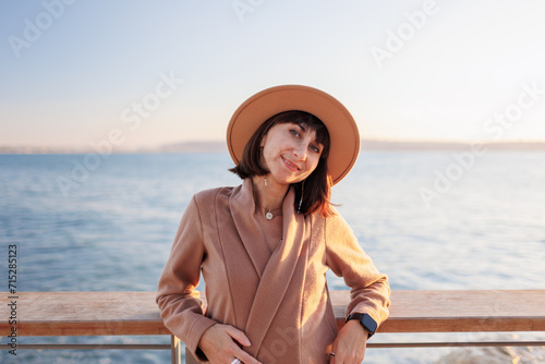 Portrait of a charming young woman in a coat and hat looking at the sea. A girl stands on the embankment and looks at the sea.