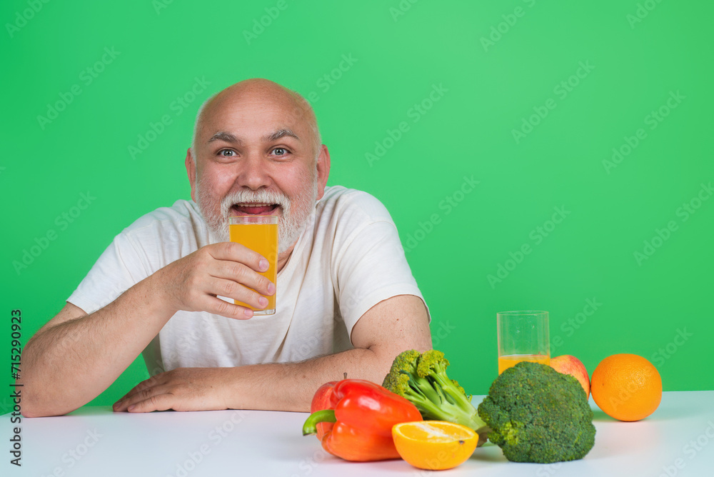 Senior drink orange juice near vegetables in studio. Elderly man hold glass of orange juice and vegetables. Orange juice and vegetables. Fruit and vegetables for aged people. Healthy lifestyle Senior drink orange juice near vegetables in studio. Elderly man hold glass of orange juice and vegetables. Orange juice and vegetables. Fruit and vegetables for aged people. Healthy lifestyle