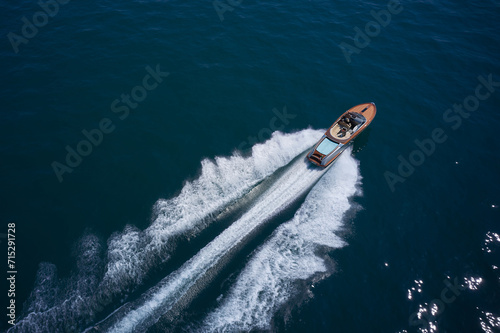 Expensive wooden boat, man and woman in motion on the water making a white trail looking like air. A large modern high-speed wooden luxury boat moves on blue water, top view.