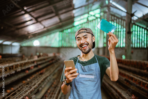 Smiling Asian businessman showing a bankbook and holding a phone standing at a chicken farm