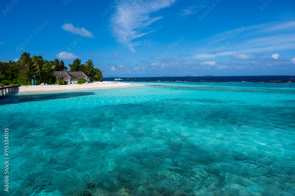 Fototapeta premium View of the lagoon with pale blue water and the ocean in the background. On the left side is an island with a sandy beach, palm trees and tropical houses.
