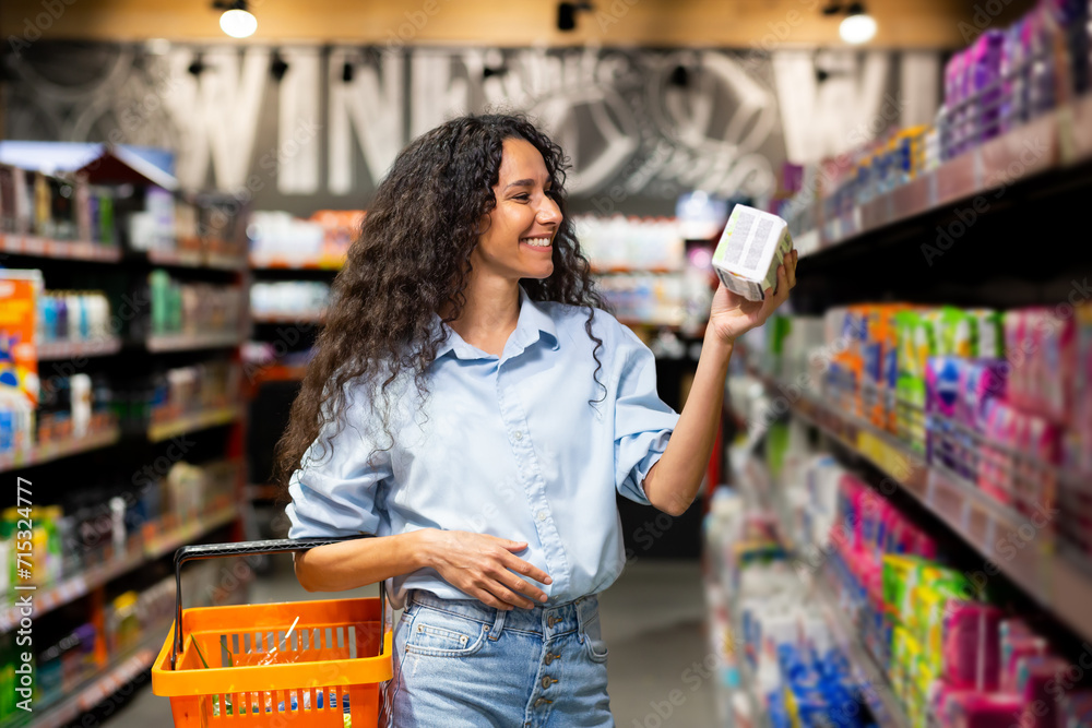 Joyful young woman with curly hair selects products in supermarket, exemplifying healthy lifestyle and shopping habits.