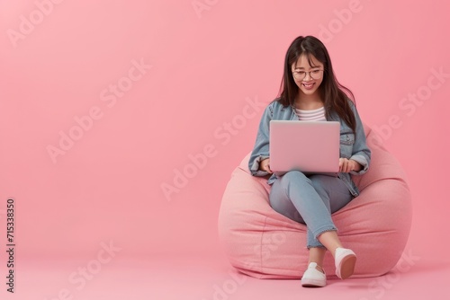 Full body young happy young woman of Asian ethnicity sit in bag chair use work on laptop isolated on plain pink background. AI generative