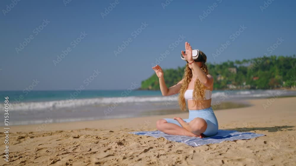 Woman meditates with VR headset on beach, virtual reality transports to tranquil space for mindfulness. Female in yoga pose, finds peace on sandy shore, waves in background. Slow motion.