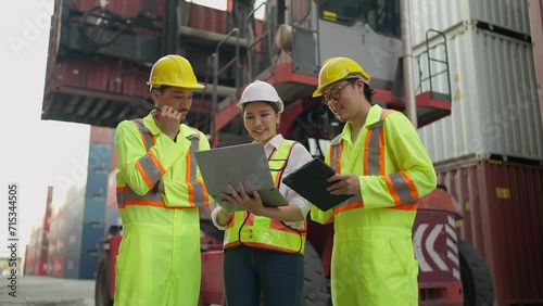 Hardworking team of workers in an industrial warehouse, wearing safety helmets working on laptop and tablet focused discusstion and talking together at cargo container yard. Teamwork concept photo