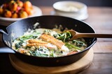 a skillet with spinach and feta stuffed chicken in a creamy sauce, wooden spatula on the side