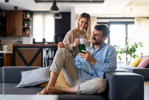 Young happy couple using phone to share social media news at home, doing shopping online.