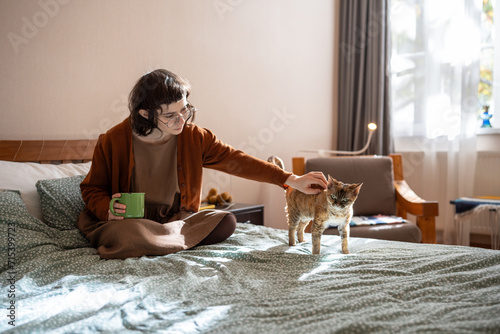 Girl petting Devon Rex cat sitting in bed at home drinking coffee at home. Woman stroking domestic pet. Spending morning time with animal starting new day. Life of lonely introverted girl with cat 