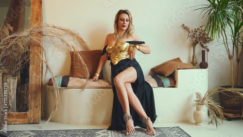 fantasy sexy woman sitting posing in room interior turkish style sofa pillows carpets. clothes gold accessories beauty face blonde hair. fashion model girl queen in black long dress golden corset photo