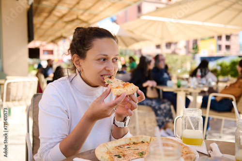 Young multiracial woman has lunch in an Italian restaurant on the summer terrace, biting a slice of delicious pizza