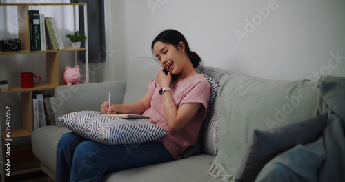 Portrait of Young woman talking mobile phone and take notes in a notebook while sitting on sofa at home office.