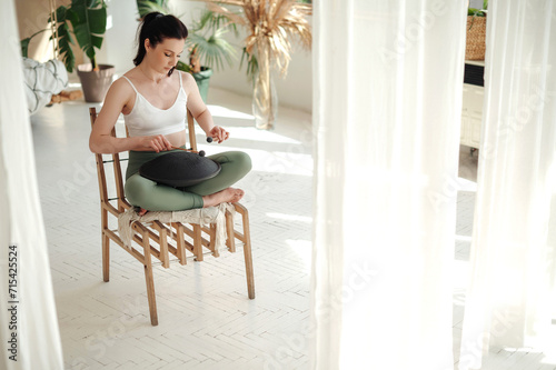 Fit brunette woman sitting on chair in lotus pose and playing on steel tongue drum. Adorable female relaxing to calm and spiritual. Concept of peace and inspiration.