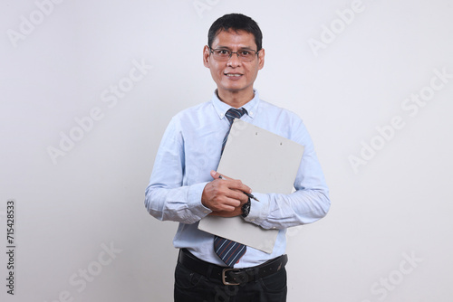 Portrait of successful Asian businessman in formal shirt and glasses looking at camera confidently while holding clipboard