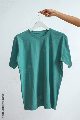 Hand holding tosca color t-shirt hanging on white cloth hanger over white background