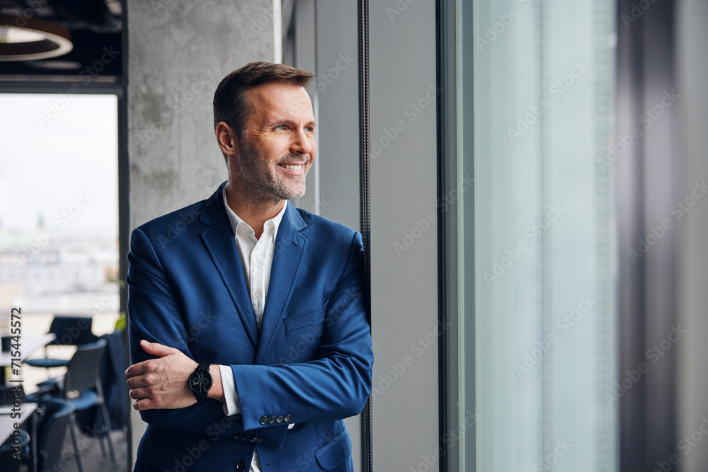 Smiling businessman standing with arms crossed near window in office Smiling businessman standing with arms crossed near window in office