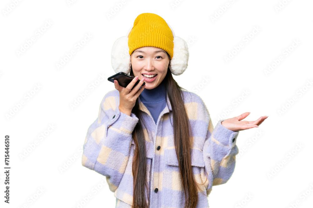 Young Asian woman wearing winter muffs over isolated chroma key background keeping a conversation with the mobile phone with someone Young Asian woman wearing winter muffs over isolated chroma key background keeping a conversation with the mobile phone with someone