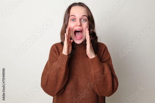 Amazed Caucasian woman wearing brown jumper standing isolated over gray background screaming with hands near mouth making advertisement yelling loud announcing good important news