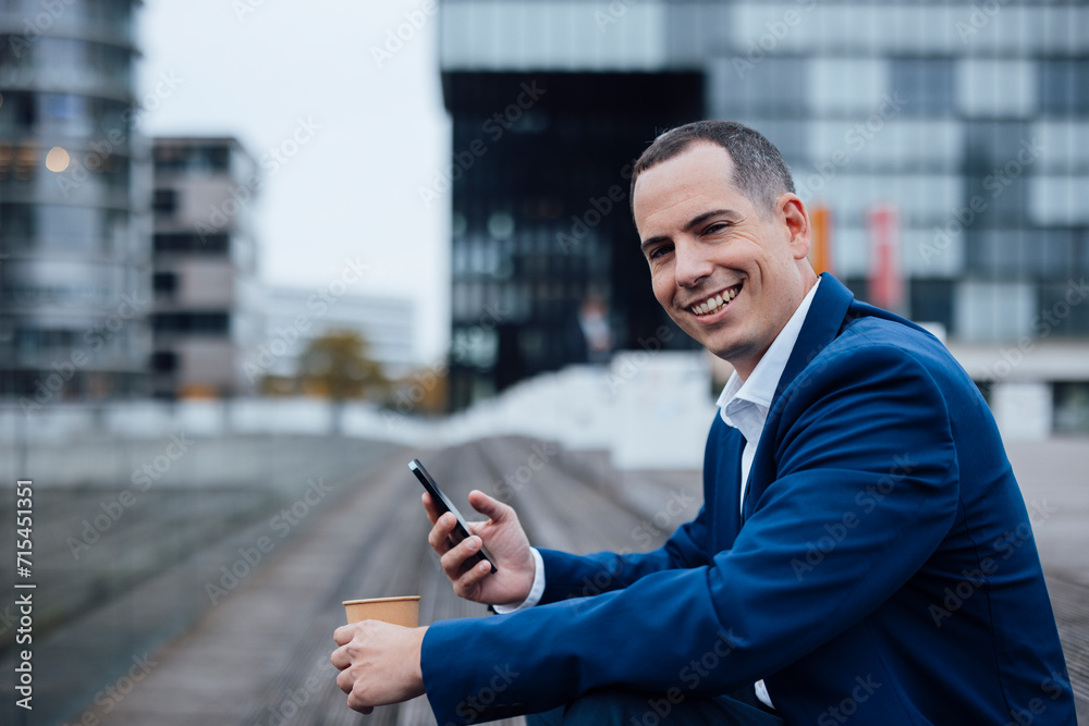 Happy businessman standing with smart phone and coffee cup in front of buildings Happy businessman standing with smart phone and coffee cup in front of buildings