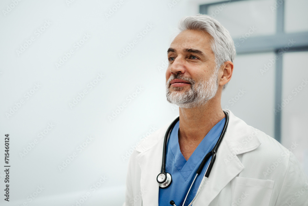 Portrait of confident mature doctor standing in Hospital corridor. Handsome doctor with gray hair wearing white coat, stethoscope around neck standing in modern private clinic, looking at camera. Portrait of confident mature doctor standing in Hospital corridor. Handsome doctor with gray hair wearing white coat, stethoscope around neck standing in modern private clinic, looking at camera.