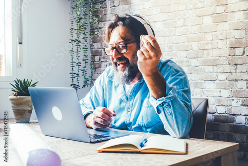 Cheerful Italian freelance man singing and listening to music with headphones while working with laptop at home. He keeps the rhythm with his hands, enjoying his favorite song.