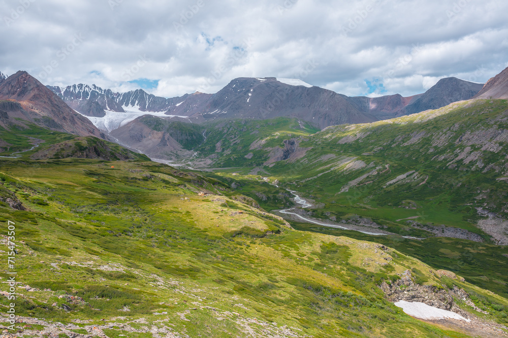 Wide green alpine valley with snake river among hills and rocks with vast view to large snow mountain top and big glacier tongue under cloudy sky. Serpentine river in high mountains. Shadows of clouds Wide green alpine valley with snake river among hills and rocks with vast view to large snow mountain top and big glacier tongue under cloudy sky. Serpentine river in high mountains. Shadows of clouds
