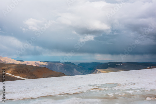 Dramatic alpine top view from flat sloping glacier on precipice edge to deep gorge between colorful sunlit cliffs and mountain range silhouette in rain under lead gray cloudy sky. Rainy grey clouds.