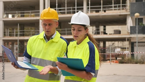 European man and woman engineers standing in building yard and discussing project documentation of new many-storeyed house. 