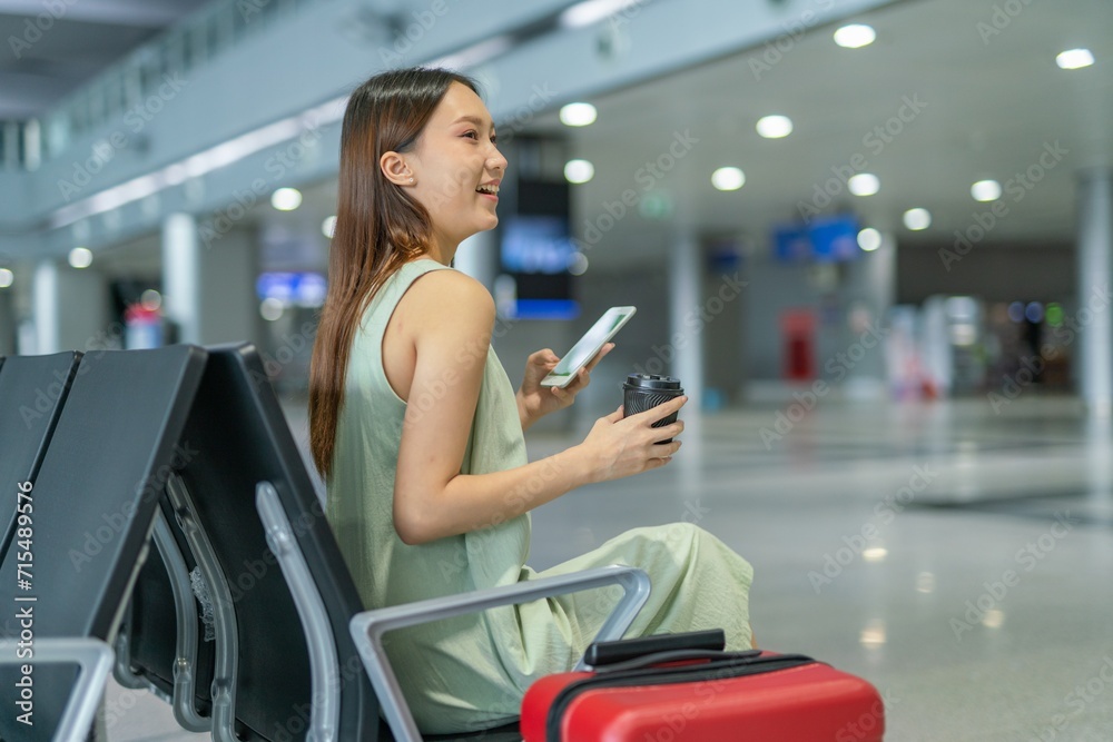Asian woman with luggage using phone while sipping a cup of coffee at a transportation departure hall