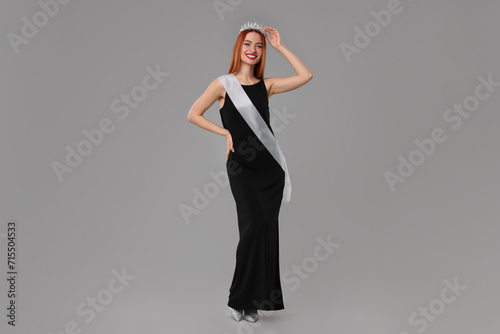 Beautiful young woman with tiara and ribbon in dress on light grey background. Beauty contest