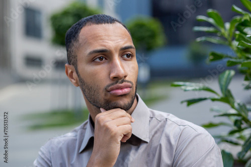 Pensive young African American man in shirt with a thoughtful expression walking near an office building or campus. © Liubomir