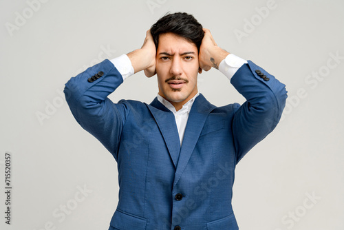 Businessman in a blue suit shows a moment of stress or confusion, gripping his head with both hands, against a plain background, illustrating the challenges and pressures faced in the corporate world