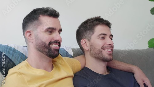 Gay Couple Watches TV together while Sitting on a Couch in the Living Room. Homossexual couple embrace, cuddle, talk, smile and watch Television Streaming Services. 
 photo