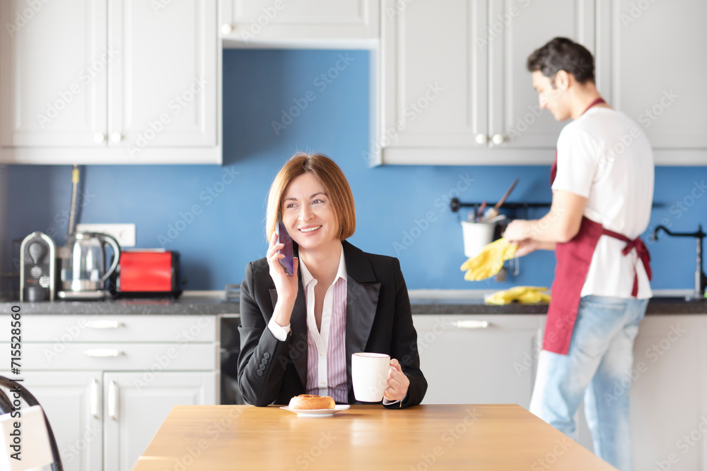 Confident business woman sits at the table and looking at her smartphone and her husband householder takes care about the house