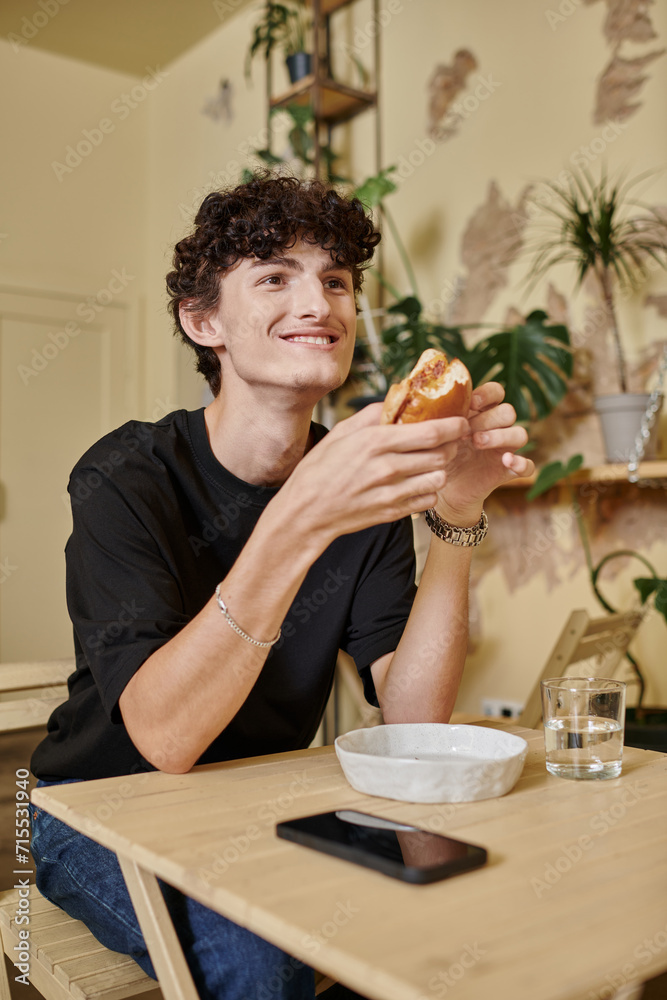 positive and curly young man holding organic tofu burger and smiling in plant filled vegan cafe positive and curly young man holding organic tofu burger and smiling in plant filled vegan cafe