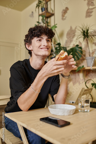 positive and curly young man holding organic tofu burger and smiling in plant filled vegan cafe