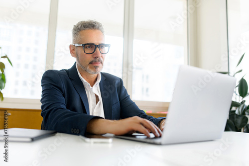 Focused mature senior businessman wearing eyeglasses using laptop in contemporary office space, concentrated 60s man employee typing, messaging, programmer develops software, looks at computer screen