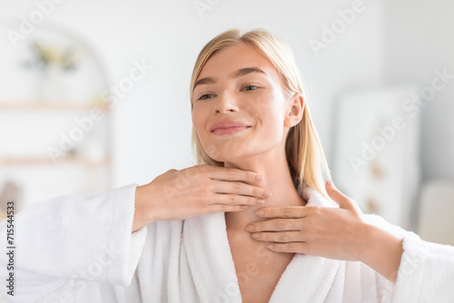 Pretty blonde woman applying moisturizing cream on her neck indoors photo