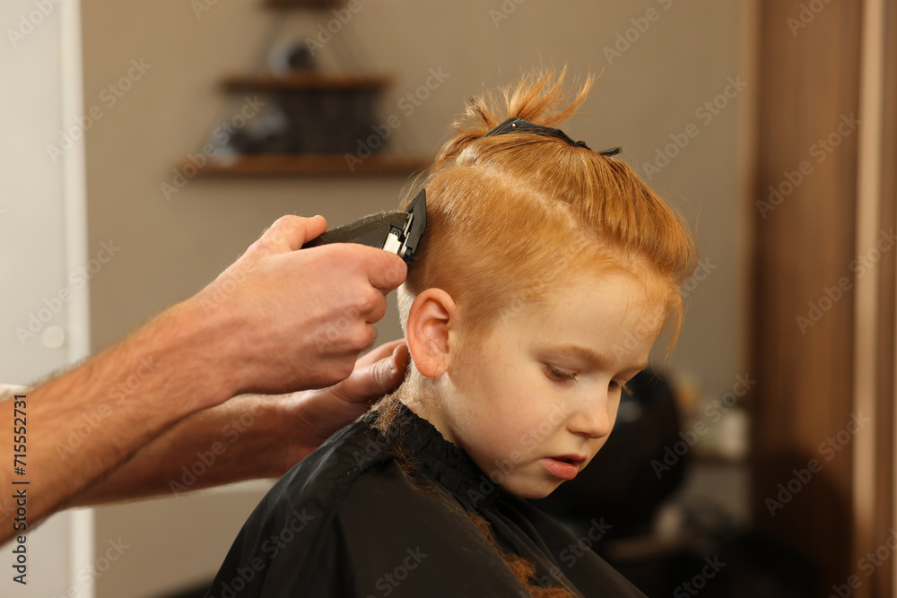 Fototapeta premium Professional hairdresser cutting boy's hair in beauty salon, closeup