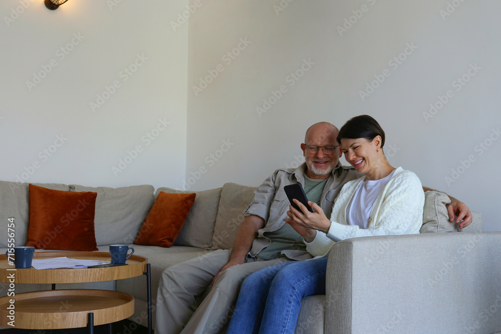 Obraz premium Adult married couple at home, sitting on the couch laughing at memes on the phone. Close up, copy space, background