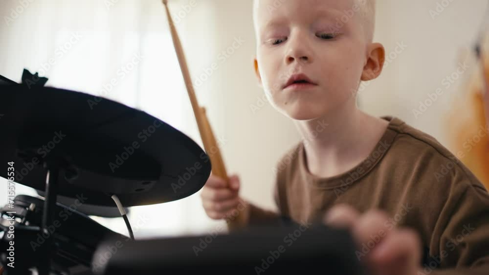 Close-up shot from below of a happy blond albino boy with white hair setting up an electronic drum kit before starting to play it with wooden sticks in his room, developing his talent and hobby