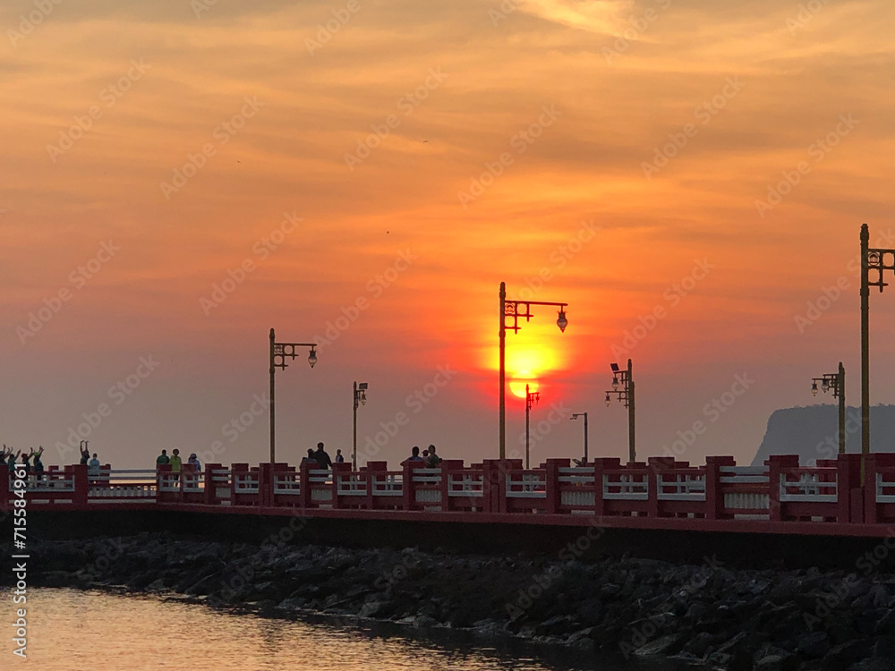 Cityscape Embraced by the Warm Glow of Sunset with Silhouetted Towers, Industrial Structures, and a Serene Pier by the Water, as the Sky Paints a Palette of Orange Hues in the Early Evening Light