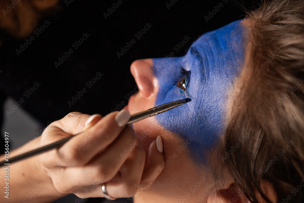 Close-up portrait of woman having her face painted blue by an artist with brush. Creative makeup, performance makeup - obrazy, fototapety, plakaty 